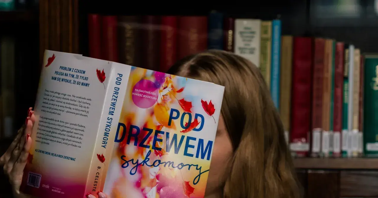 Dziewczyna czyta książkę "Pod drzewem sykomory", ukrywając twarz za okładką. W tle widać regały pełne książek.