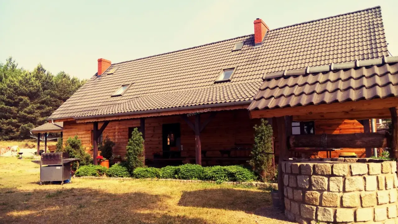 Drewniany dom pod kasztanem, z kamienną studnią i grillem, otoczony zielenią i lasem.