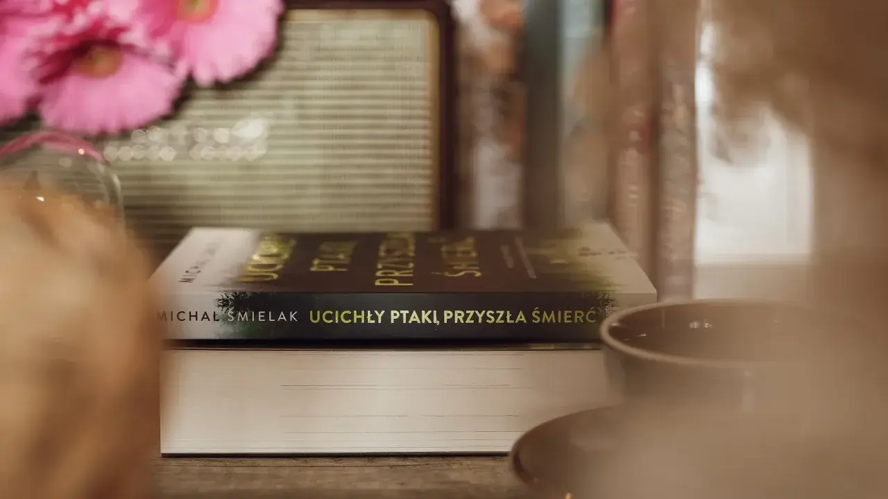 Na stole leżą książki, jedna z nich ma tytuł "Ucichły ptaki, przyszła śmierć". W tle różowe kwiaty i filiżanka.