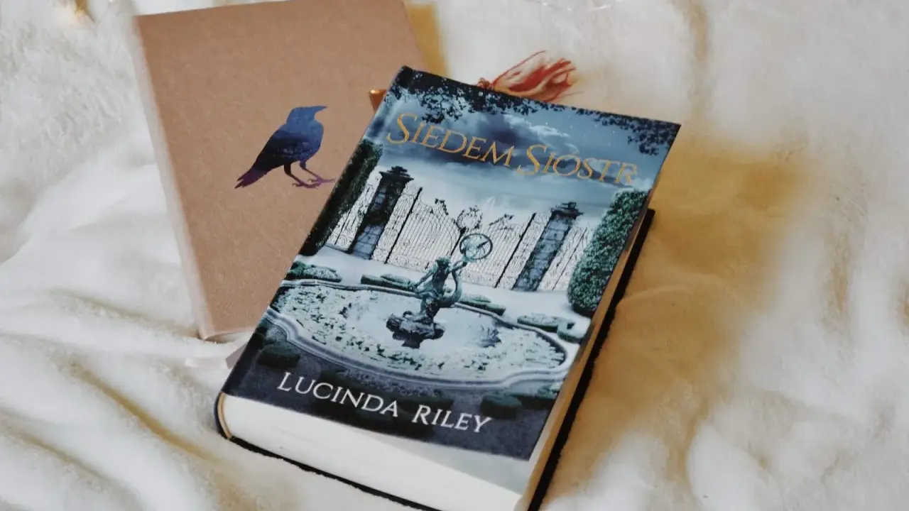 Okładka książki "Siedem Sióstr" Lucindy Riley na białym tle. Obok leży brązowy notes z wizerunkiem kruka.