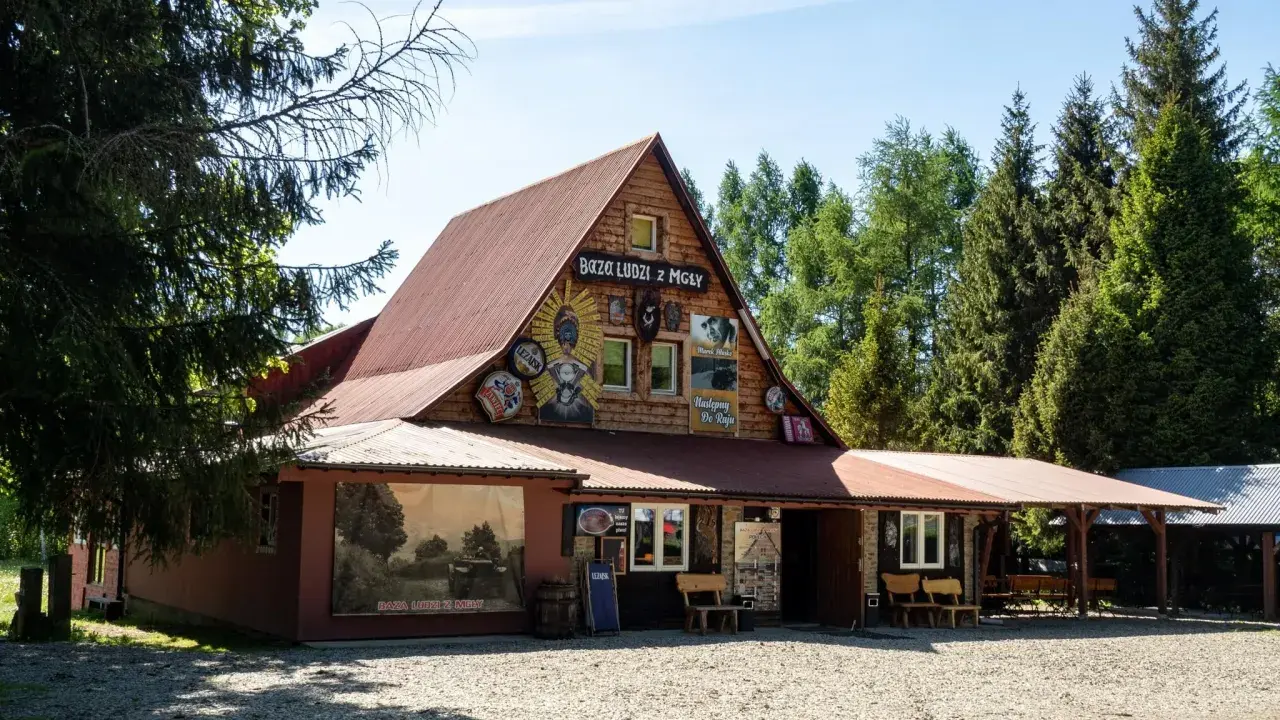 Drewniany budynek z napisem "Baza Ludzi z Mgły" i malowidłami. Obok duży mural z krajobrazem i napisem "Baza Ludzi z Mgły".