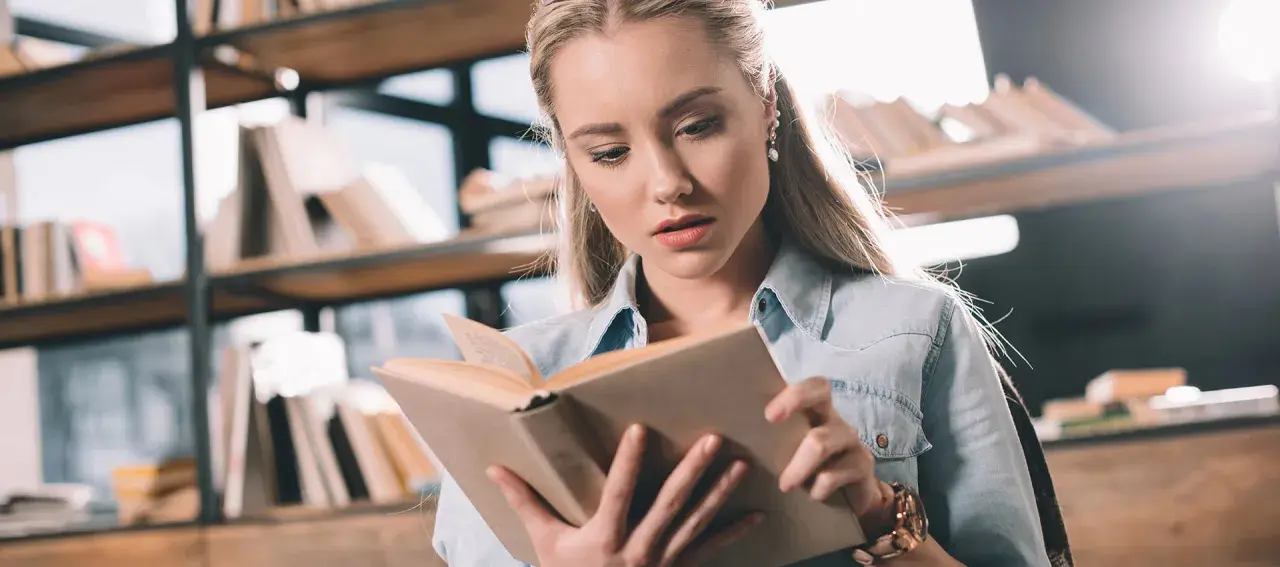 Młoda kobieta z uwagą czyta książkę po angielsku, siedząc wśród regałów pełnych literatury.