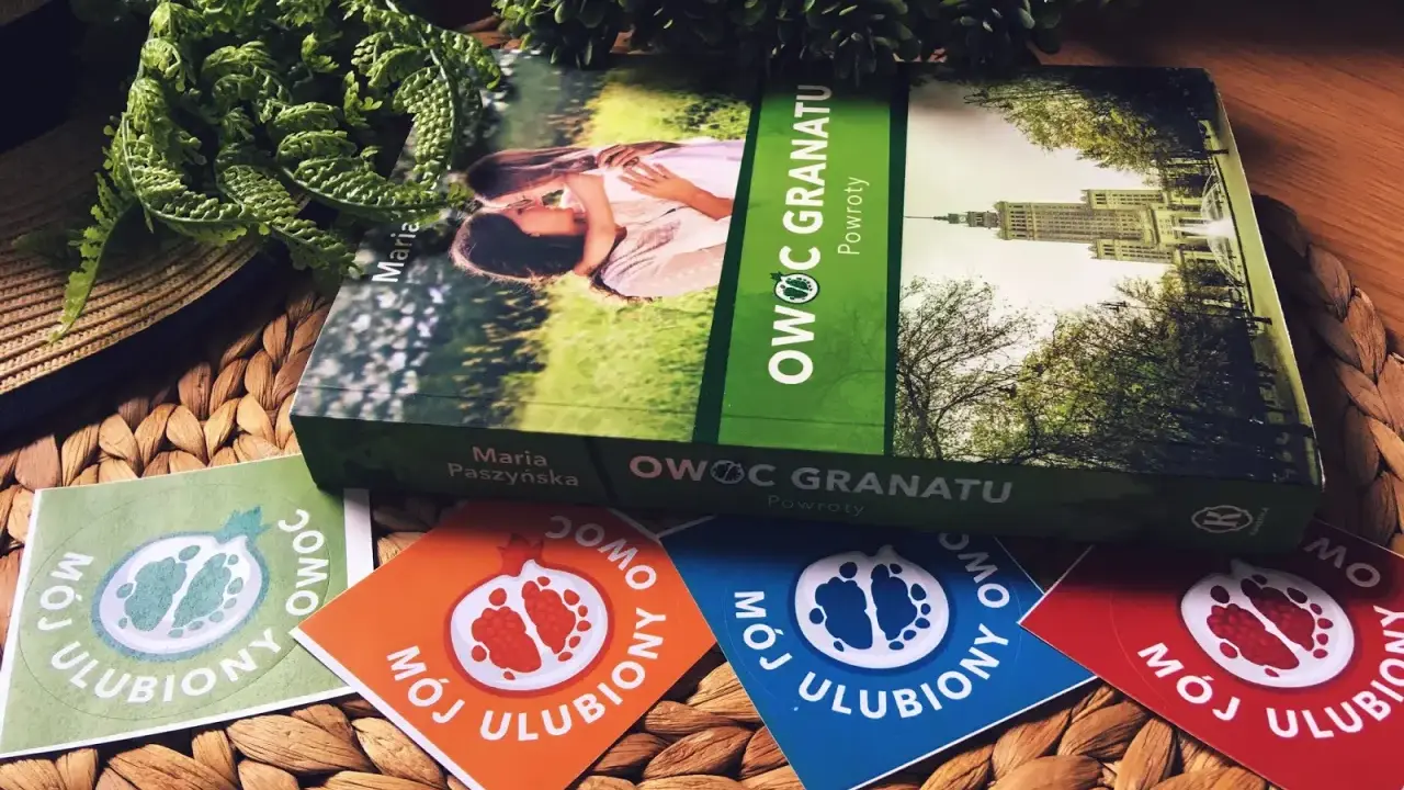 Książka "Owoc granatu" Marii Paszyńskiej z motywem pary na okładce, otoczona zielenią i kolorowymi naklejkami z symbolem granatu.
