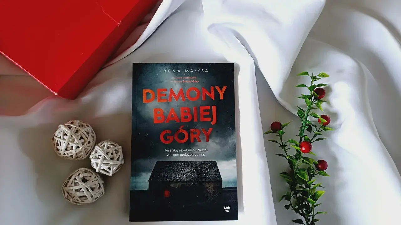Książka "Demony Babiej Góry" Ireny Małysy, leżąca na białym jedwabiu, obok czerwonego pudełka i ozdobnych kulek. Historia, która zabiera daleko od Babiej Góry.