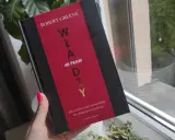 Dłoń z neonowym różowym lakierem trzyma książkę "48 praw władzy" Roberta Greene'a.