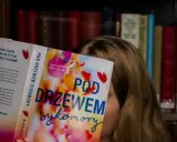 Dziewczyna czyta książkę "Pod drzewem sykomory", ukrywając twarz za okładką. W tle widać regały pełne książek.