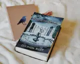 Okładka książki "Siedem Sióstr" Lucindy Riley na białym tle. Obok leży brązowy notes z wizerunkiem kruka.