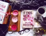 Książka "It Ends With Us" Colleen Hoover, obok świec, kawy i innych książek, tworzy przytulną atmosferę do czytania.