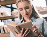 Młoda kobieta z uwagą czyta książkę po angielsku, siedząc wśród regałów pełnych literatury.