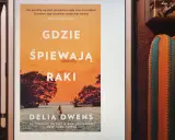 Dziewczyna z bagien na okładce książki "Gdzie śpiewają raki". Samotna postać w kajaku na tle zachodzącego słońca.