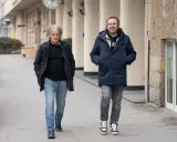 Jacek Podsiadło, ojciec Dawida, idzie ulicą w granatowej kurtce i jeansach. Obok niego młodszy mężczyzna w podobnym stroju.