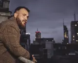 Chmielarz długa noc. Mężczyzna w płaszczu w kratę spogląda na nocne miasto z dachu.