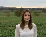 Kobieta w białej bluzce z koronką stoi na polu. W tle widać pagórki i drzewa. Czuje, że nie to miejsce, nie ten czas.
