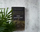 Okładka książki Kristin Hannah "Gdzie poniesie wiatr" na tle szarej ściany, z zielonymi liśćmi rośliny w zbliżeniu.