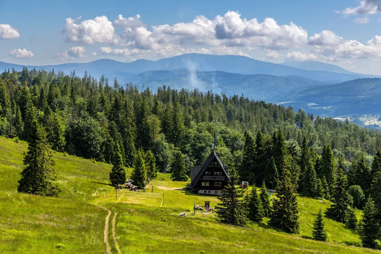 Drewniany domek w górach, otoczony lasem, z dymem unoszącym się z komina. Widok na pasmo górskie daleko od Babiej Góry.